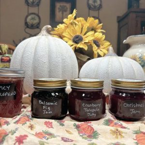 a photo of four jars of products in a fall decorated setting: spicy jalapeño jelly, balsamic fig jam, cranberry jalapeño jelly, and Christmas jam.