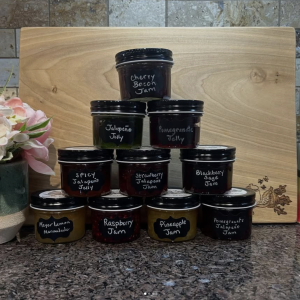 A photo on 10 jars of jellies and jams: cherry bacon jam, jalepeno jelly, pomegranate jelly, spicy jalapeño jelly, strawberry jalapeño jam, blackberry sage jam, Meyer lemon marmalade, raspberry jam, pineapple jam, and pomegranate jalapeño jam.