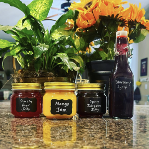 a photo of four jars on a counter in front of some flowers: prickly pear jelly, mango jam, spicy jalapeño jelly, and blueberry syrup