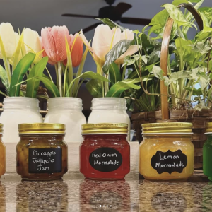 A photo of five jars of products from the farm in front of flowers: orange marmalade, pineapple jalapeño jam, red onion marmalade, lemon marmalade, and mint jelly